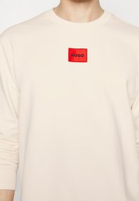 Sudadera de color beige claro con cuello redondo, puños acanalados y un parche rectangular rojo con el texto negro "HUGO" en el centro. Textura de tela suave.