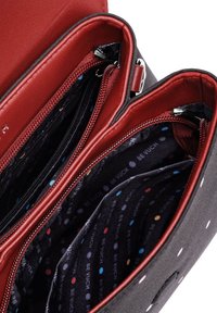 Černá textilní taška s červenými koženými akcenty, vybavená několika zipovými kapsami, podšívkou s puntíkovaným vzorem a kovovými detaily.