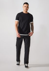 Schwarzes geripptes T-Shirt mit weißen Besätzen an den Ärmeln, kombiniert mit schwarzen Cargo-Hosen mit Seitentaschen. Modell trägt schwarz-weiße Sneakers.