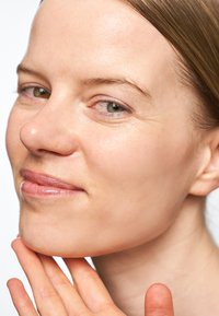 Nærbilde av et ansikt med lys hud, lysebrunt hår og en jevn tekstur. Personen hviler hånden på haken, noe som viser subtile ansiktskonturer.