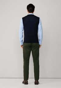 Námornícky bezrukávový sveter cez svetlomodrú dlhú košeľu, kombinovaný s tmavozelenými nohavicami a hnedými topánkami, zobrazený zozadu.