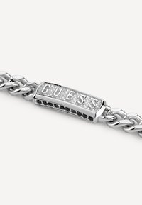 Bracciale argento con design a catena collegata e un pendente rettangolare con la scritta "GUESS" incisa, accentuato da pietre chiare e nere.