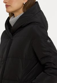 Crna puf jakna s kapuljačom, s teksturiranim sivim rebrastim ovratnikom i malim logotipom na rukavu. Glatka, kviltana površina.