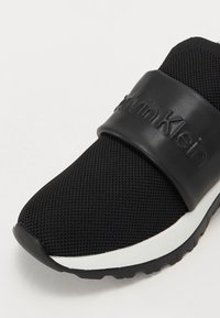Musta urheilukengät, joissa on verkkokankainen päällinen, suuri musta kumihihna, jossa on "Calvin Klein" -logo, ja valkoinen pohja, jossa on teksturoitu musta ulkopohja.