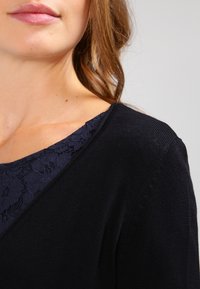 Černý svetr s hlubokým výstřihem do V, zdobený námořnicky modrou krajkou na límci. Měkká pletená textura s žebrovanými akcenty na rukávech.
