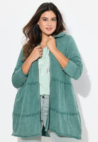 Grüne, zugeknöpfte Jacke aus weichem Stoff mit einem gestuften Design und elastischen Akzenten. Lange Ärmel und eine lässige Passform runden den Look ab.