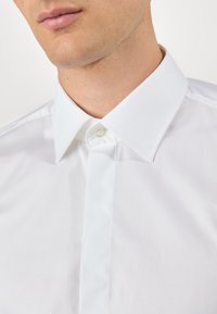 Gros plan d’un homme portant une chemise blanche à col pointu avec le bouton du haut fermé.