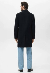 Palton din lână închisă, cu deschidere unică în spate, asortat cu blugi bleumarin și pantofi negri. Designul prezintă un guler clasic.