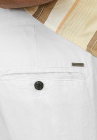 Fehér szövet rövidnadrág hátsó zsebbel, amelyen egy fekete gomb és egy ezüst címke szerepel, rajta a "JACK & JONES" felirattal. Strukturált felület és egyszerű dizájn.
