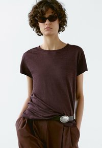 Burgundské tričko s kulatým výstřihem z měkkého materiálu, krátké rukávy, stylizované s tmavohnědými kalhotami s vysokým pasem a širokým opaskem se stříbrnou sponou.