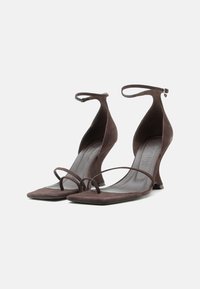 Sandalias de tacón alto de ante marrón con tiras en el tobillo, tiras finas en los dedos y tacones de aguja curvados de forma única sobre un fondo blanco.