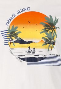 Grafiek op een wit T-shirt toont een oranje zonsondergang, silhouetten van twee paddleboarders, palmbomen en tekst met de woorden "PARADIJS UITSTAPJE."