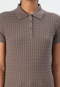Braunes, kabelstrickartiges Poloshirt mit kurzen Ärmeln, einem Kragen und einer Knopfleiste mit drei Knöpfen; mit einem kleinen Markenlogo auf der linken Brust.