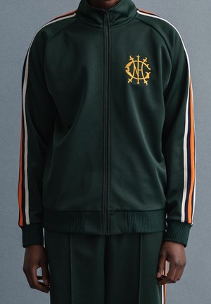 Chaqueta deportiva verde hecha de un tejido suave, con un logotipo dorado en el pecho y franjas contrastantes en naranja, blanco y azul marino a lo largo de las mangas.