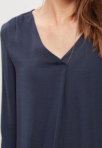 Mörkblå långärmad blus med V-ringning, tillverkad av ett mjukt tyg. Har rynkade axlar och en avslappnad passform.