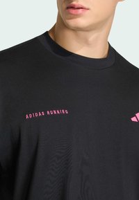 Μαύρο αθλητικό μπλουζάκι Adidas με στρογγυλή λαιμόκοψη, που φέρει ροζ λογότυπο "ADIDAS RUNNING" και μικρό ροζ λογότυπο-έμφαση στο μανίκι.