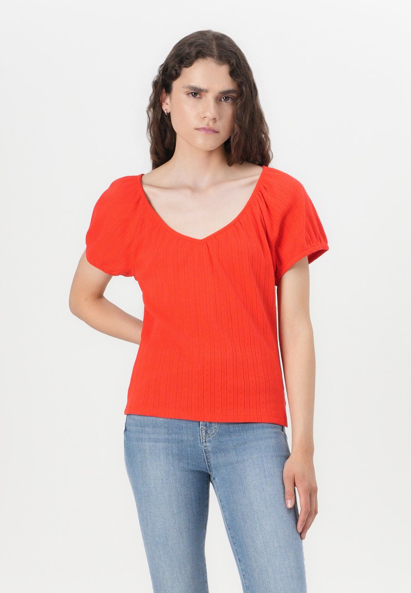 Rotes, geripptes Strickoberteil mit V-Ausschnitt und Puffärmeln. Glatte Textur mit dezenten vertikalen Linien, kombiniert mit hellblauen Jeans.