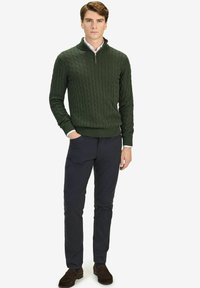 Donkergroen kabelgebreid sweater met een kwart-rits kraag, gecombineerd met donkere broek en bruine schoenen, staand tegen een witte achtergrond.