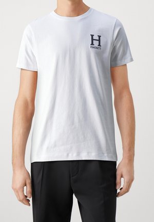 Biała bawełniana koszulka z krótkimi rękawami, okrągłym dekoltem i granatowym haftowanym logo na lewej piersi; zestawiona z czarnymi spodniami.