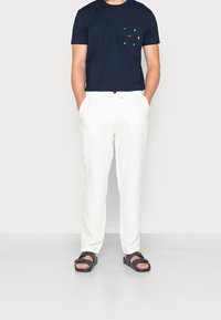 Mörkblå bomullströja med ett mönstrat ficka, i kombination med vita linbyxor och svarta sandaler med spänningsdetalj.