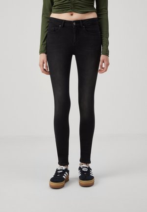 Personne portant un jean skinny noir, un haut vert à manches longues froncées et des baskets Adidas noires avec des bandes blanches et des semelles marron.