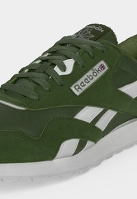 Πράσινο και λευκό παπούτσι Reebok με λεπτομέρειες σουέτ, κορδόνια και ετικέτα λογότυπου με τη σημαία του Ηνωμένου Βασιλείου στο πλάι.