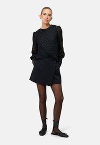 Fekete csipke blúz texturált, felfújt ujjakkal, fekete, gombos részletű átkötős szoknyával párosítva. A megjelenést fekete harisnya és lapos cipő egészíti ki.