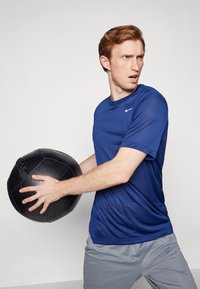 T-shirt de sport bleu, à manches courtes, logo Nike. Shorts gris clair. Individu tenant un ballon médicinal noir, expression concentrée, fond uni.