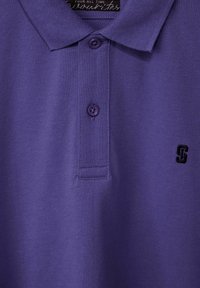 Polo shirt violet en tissu texturé, avec trois boutons au col et un petit logo noir brodé sur le côté gauche.