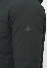 Fekete steppelt dzseki sima textúrával, kapucnival és a bal ujjon található kör alakú, tonális logóval. Minimalista design elemek.