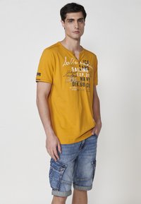 Geltona trumpa rankovės T-marškiniai su tekstu ir grafika, derinami su šviesiai mėlynomis džinsinėmis šortais, kuriuose yra cargo kišenės ir apvynioti kraštai.