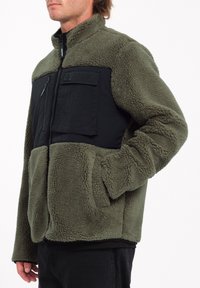 Chaqueta de forro polar en verde oliva con un panel de nylon negro y un bolsillo en el pecho. Cuenta con cuello alto y dos bolsillos frontales. Textura suave.