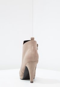 Bota de tacón alto de ante beige con cremallera en la parte posterior, de pie sobre una superficie blanca contra un fondo blanco oscuro.