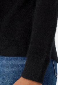 Svart, fluffig tröja med ribbad manschettdetalj, kombinerad med blå denim. Tyget verkar mjukt och texturerat och visar upp en close-fitting design.