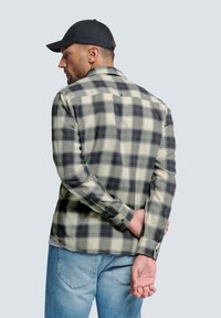 Karierte grüne und beige Hemd aus Stoff, mit langen Ärmeln und Knopfverschluss, getragen mit hellblauen Jeans und einer schwarzen Kappe.