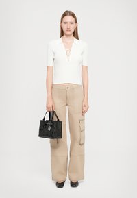 Top corto a maniche corte bianco con dettaglio in pizzo, abbinato a pantaloni cargo beige. Borsa a tracolla nera con motivo e scarpe nere senza lacci.