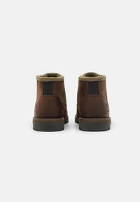 Botas de cuero marrón con un suave cuello de forro polar verde y paneles elásticos negros en los costados. Suela de goma resistente con dibujo visible.