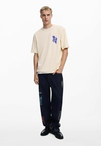 Cream-colored oversized T-shirt with blue graphic detail, paired with dark denim pants featuring multicolored patches and a loose fit.