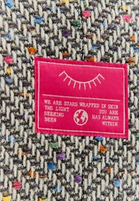 Textilní štítek na vícebarevné tkanině; fialové, modré a oranžové skvrny na šedém pozadí; štítek s růžovou látkou obsahuje bílý text.