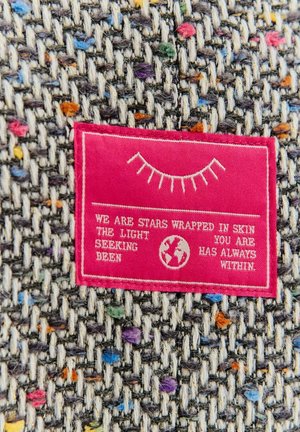 Etykieta tekstylna na wielokolorowej tkaninie tkanej; fioletowe, niebieskie, pomarańczowe plamki na szarym tle; etykieta z różowego materiału z białym tekstem.