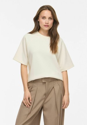 Giovane donna con lunghi capelli castani che indossa una maglia corta a maniche corte color crema e pantaloni beige plissettati a vita alta, in piedi davanti a uno sfondo bianco.