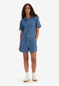 Dżinsowy kombinezon krótki z krótkimi rękawami, z przodem na guziki oraz kieszonką na piersi. Jasnoniebieski kolor z widocznymi detalami szycia i luźnym krojem.