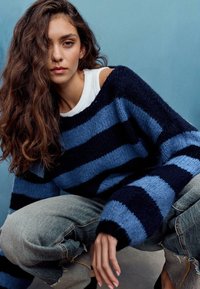 Gestrickter Pullover mit breiten horizontalen blauen und marineblauen Streifen, locker sitzend und mit übergroßen Ärmeln, kombiniert mit ausgewaschenen hellen Jeans.