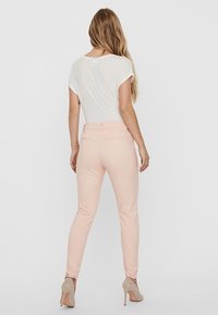 Žena stojící zády, oblečená v bílém tričku s krátkým rukávem, světle růžových slim-fit kalhotách a béžových lodičkách na jednobarevném pozadí.