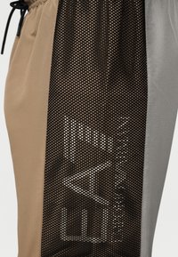Gros plan sur un pantalon beige et gris avec un panneau en mesh noir affichant un logo géométrique blanc sur le côté.