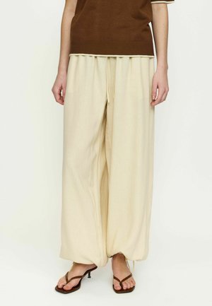 Személy laza, bő, krémszínű nadrágban, zsinórral húzott szegélyekkel, barna rövid ujjú felsőben és barna magassarkú szandálban, egyszínű háttér előtt állva.