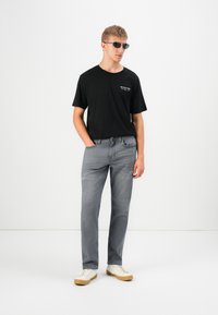 Joven vestido con una camiseta negra, jeans grises, zapatillas blancas y gafas de sol negras, de pie con las manos en los bolsillos contra un fondo blanco.