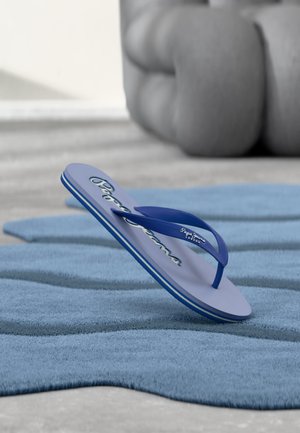 Blauer Pepe Jeans Flip-Flop, balanciert auf der Ferse auf einem strukturierten blauen Teppich mit einer verschwommenen großen grauen Skulptur im Hintergrund.