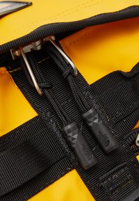 Yellow waterproof fabric with black straps and hardware; features black zip pulls with textured grip and logo, reinforced stitching visible.