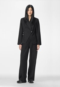 Una mujer está de pie mirando hacia adelante, vestida con un traje negro de raya diplomática de gran tamaño con pantalones de pierna ancha y zapatos negros, sobre un fondo blanco liso.